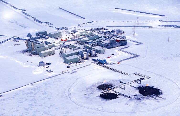 Industrial buildings in the snow have several roads and pipelines running to them and three wells with flares and blackened areas around them.