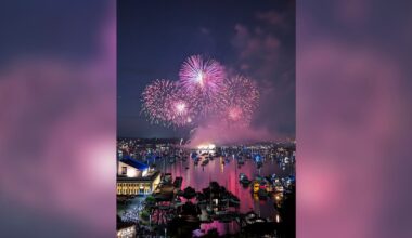 Colorful fireworks burst over a harbor at night, illuminating boats on the water and buildings along the shore. The sky is filled with pink and purple lights reflecting on the calm surface below.