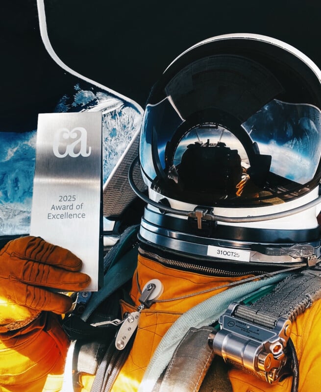 An astronaut in an orange suit and helmet holds a silver plaque that reads "2025 Award of Excellence" with the letters "ca" on top, sitting in a spacecraft with Earth visible outside the window.