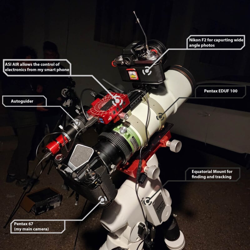 A telescope setup at night with attached equipment labeled: ASI AIR for electronic control, autoguider, Pentax 67 main camera, Nikon F2 for wide-angle photos, Pentax EDUF 100 lens, and equatorial mount for tracking.