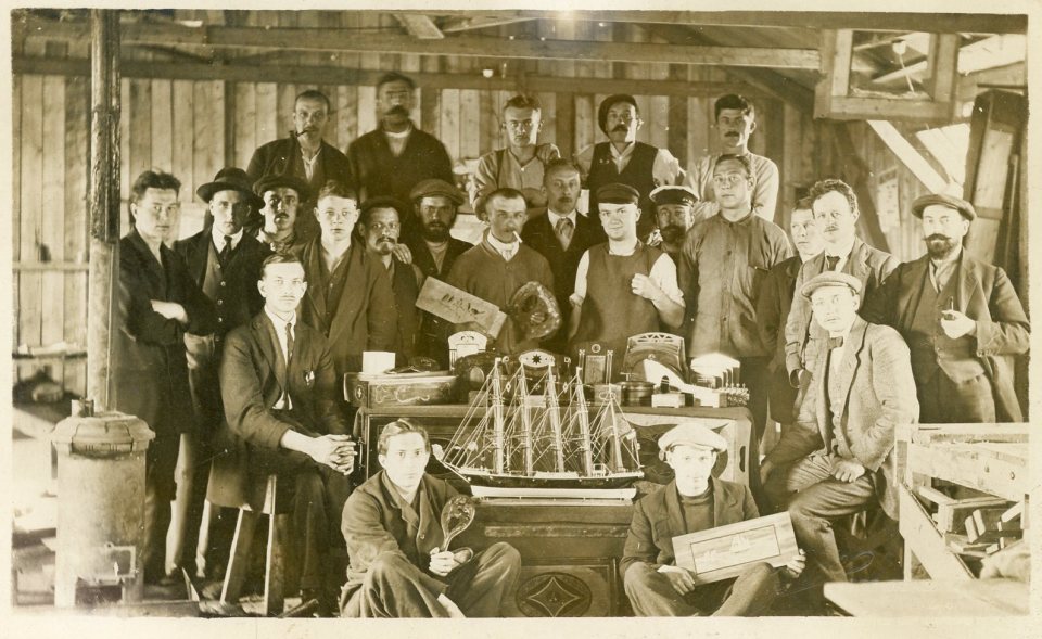Photo of a large group of men posing in a workshop with a model ship.