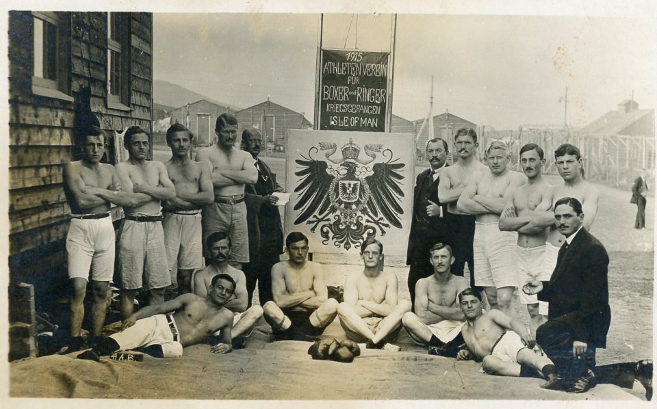 Photo of a group of shirtless men posing in front of a sign for a 1915 athletic club for boxers and wrestlers.