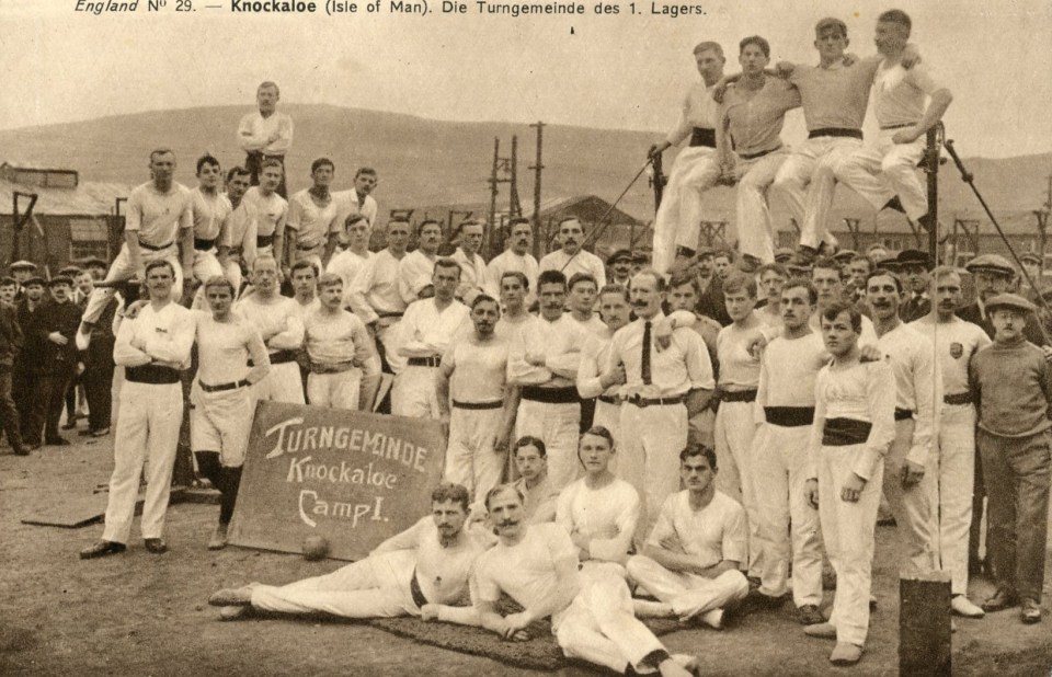 Photo of a large group of men in white uniforms, possibly a sports team, posing for a photograph in front of a sign that reads "Turngemeinde Knockaloe Camp."