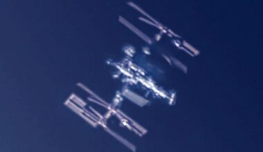 A blurred, close-up image of the International Space Station (ISS) orbiting against a dark blue sky, with its solar panels and structural modules visible.