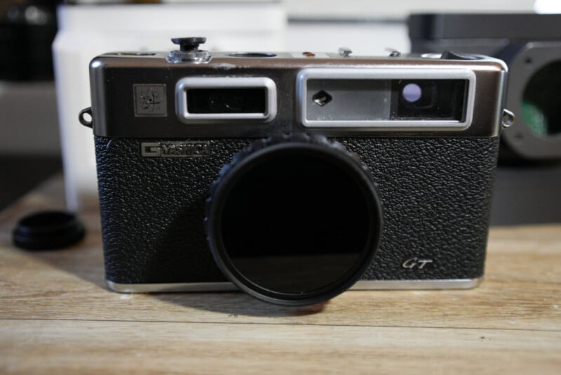 A vintage Yashica GT film camera with a black textured body sits on a wooden surface. The lens has a round filter attached, and the camera’s top controls and viewfinders are visible.