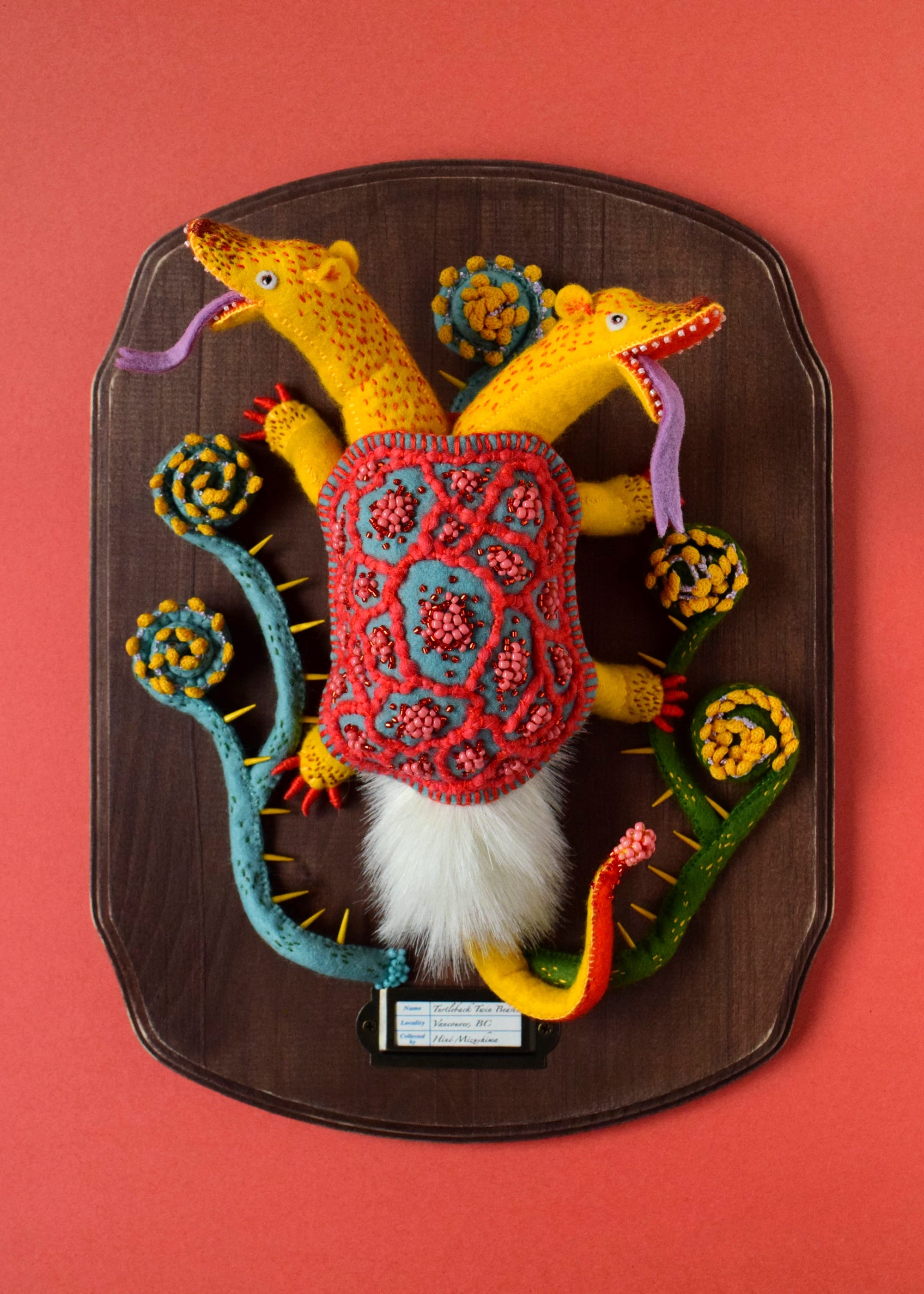 a felted sculpture of a two-headed creature with a turtle shell on its back, mounted on a wooden plaque