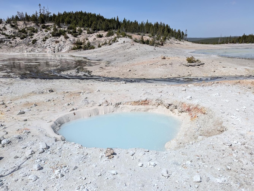 photo of a roughly round hole in the ground full of milky pale-blue water. the ground is white, with rubble strewn around. in the distance, a small hill topped with pine trees is visible under a clear blue sky.