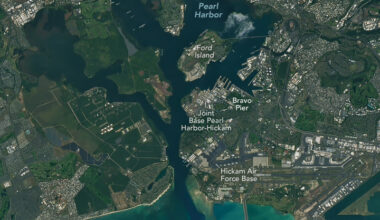 A satellite image shows Pearl Harbor in Hawaii. Several piers, runways, and large buildings are visible around the harbor — parts of Joint Base Pearl Harbor-Hickam. Bravo Pier, where the Apollo 11 astronauts docked after splashdown following their mission to the Moon, is labeled.