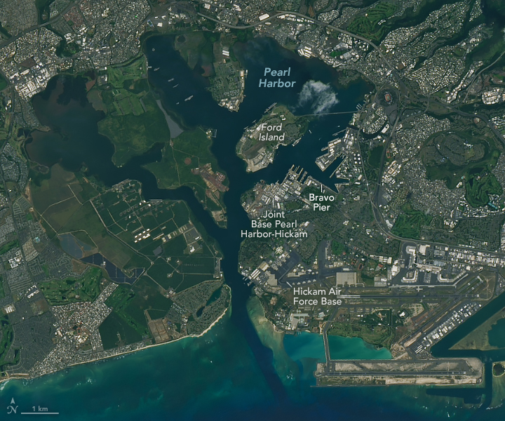 A satellite image shows Pearl Harbor in Hawaii. Several piers, runways, and large buildings are visible around the harbor — parts of Joint Base Pearl Harbor-Hickam. Bravo Pier, where the Apollo 11 astronauts docked after splashdown following their mission to the Moon, is labeled.