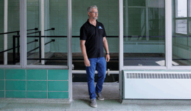 A man wearing a black polo shirt, jeans and sneakers walks through an interior doorway. He has short grey hair and a trimmed beard. He is wearing glasses. There is green tile on the wall behind him and a staircase leading down. Either side of the doorway is panelled glass.