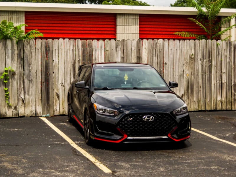 A black Hyundai Veloster N with red accents is parked in an empty parking lot in front of a weathered wooden fence and a building with red and white siding.