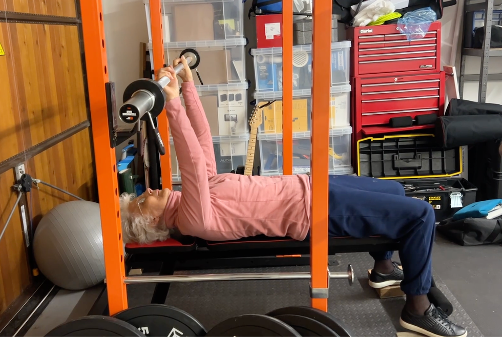 an older, white haired woman, wearing navy trousers and a pink fleece, bench pressing the bar.