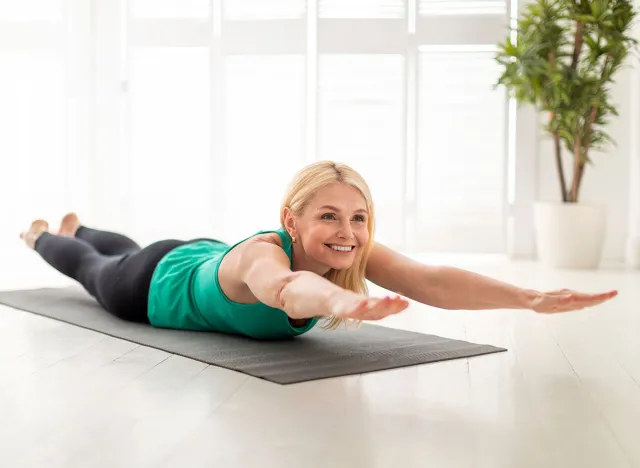 Sporty Senior Woman Making Superman Exercise While Training At Home, Smiling Mature Lady Doing Back Bending While Lying On Fitness Mat In Light Living Room, Raising Arms And Legs, Free Space