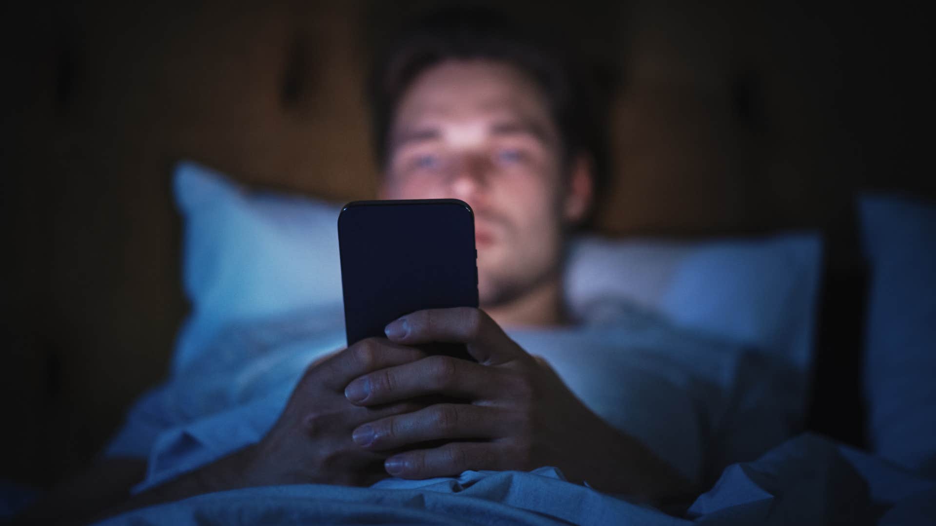 Man with a phone charger on his nightstand sitting in the dark.