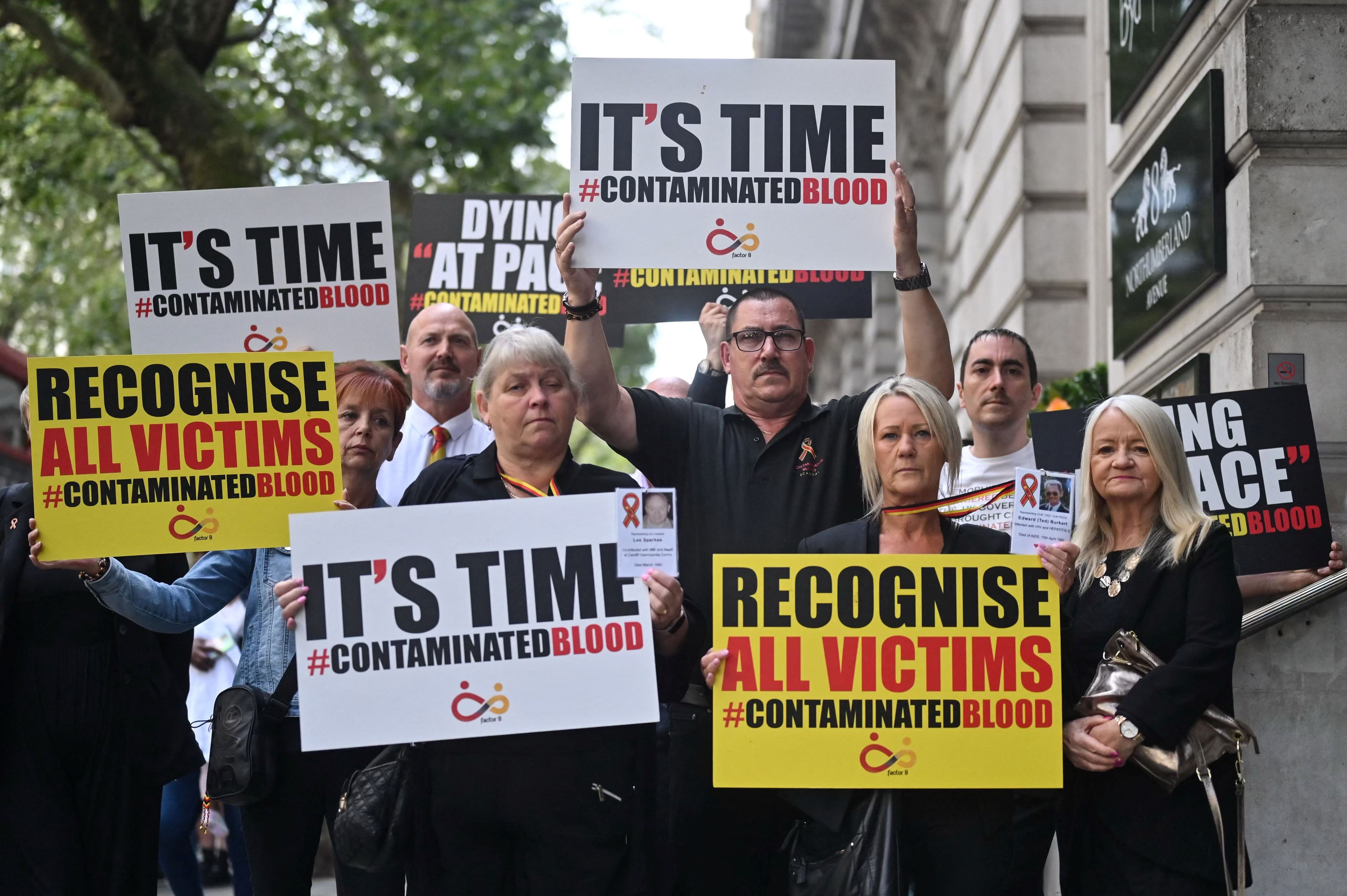 Demonstrators protesting the NHS infected blood scandal hold signs reading "It's time #ContaminatedBlood" and "Recognise all victims #ContaminatedBlood".