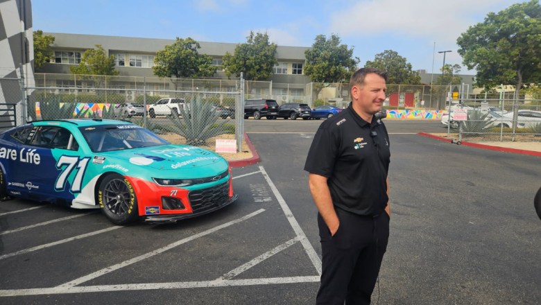 A NASCAR driver stands in a lot in front of an aqua race car.