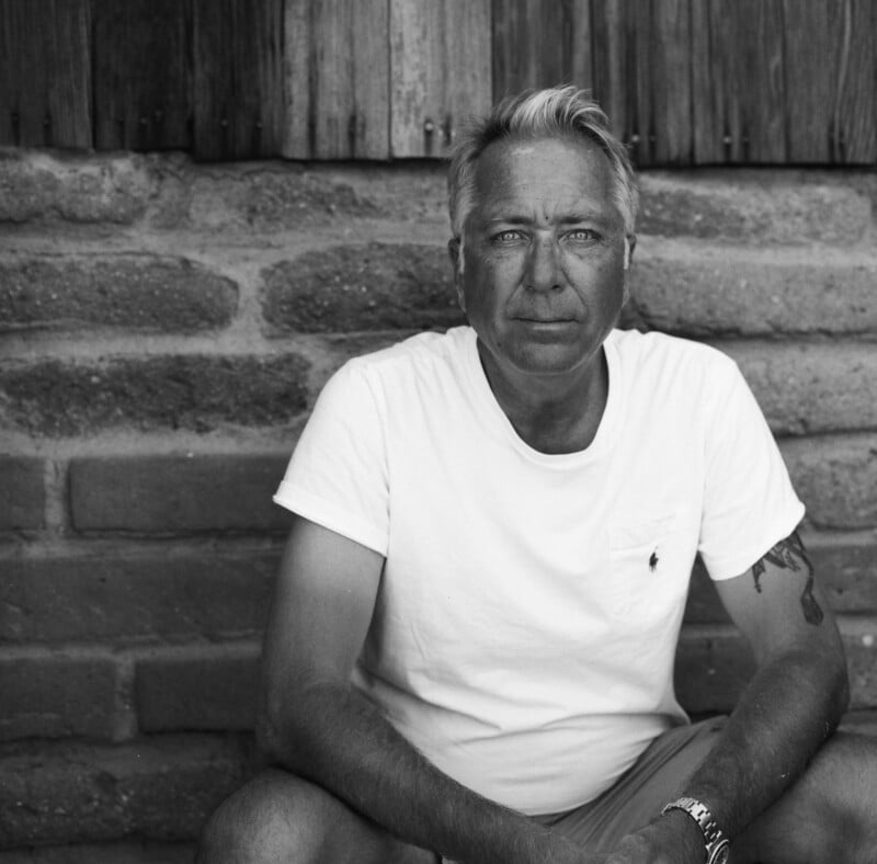 Black and white photo of a middle-aged man with light hair, sitting in front of a brick and wood wall. He wears a white t-shirt and shorts, with a tattoo on his left arm and a watch on his right wrist.