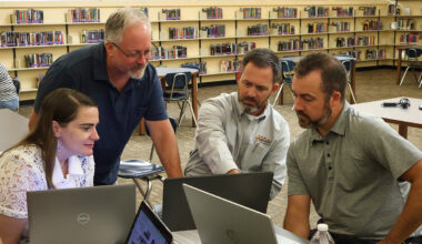 Four educators collaborate around laptops in a library during a professional development session. One person is standing and pointing at a screen while the others focus attentively, highlighting teamwork and engagement in a learning environment.