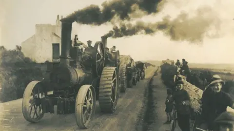 MNH Stream driven engines with plumes of smoke bellowing out of their funnels transporting the whale along a dirt road.