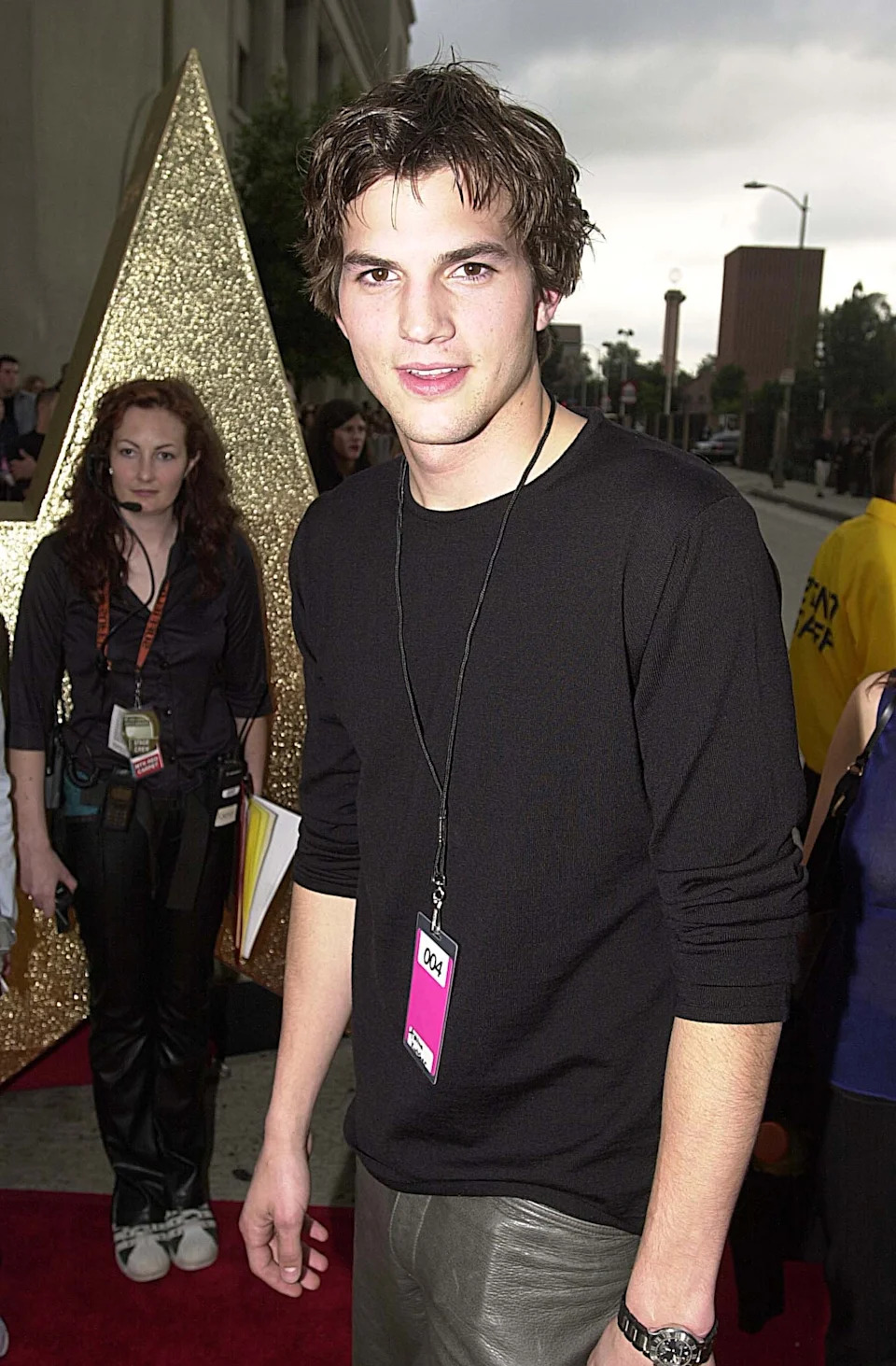 Person on a red carpet in a black long-sleeve shirt and leather pants, with a lanyard around their neck. Large gold star prop in the background