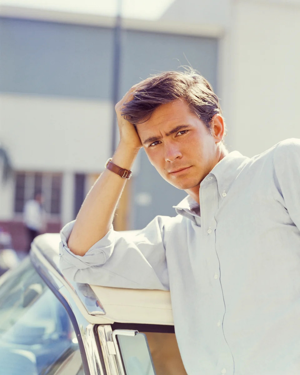 Person leaning on a car, wearing a button-up shirt, with a thoughtful expression, in a casual outdoor setting