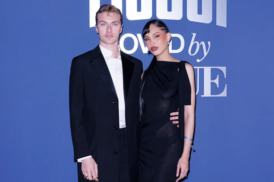 Lucky Blue Smith and Nara Smith posing together at an event, dressed in stylish formal attire. A logo is visible in the background