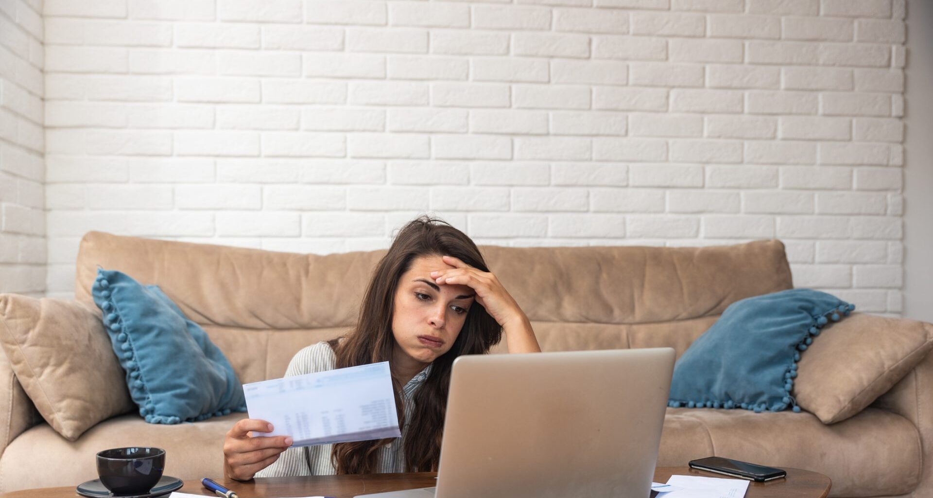 Young woman sitting on the floor in front of a couch, looking at a laptop and papers with one hand on her forehead and a stressed expression.