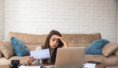 Young woman sitting on the floor in front of a couch, looking at a laptop and papers with one hand on her forehead and a stressed expression.