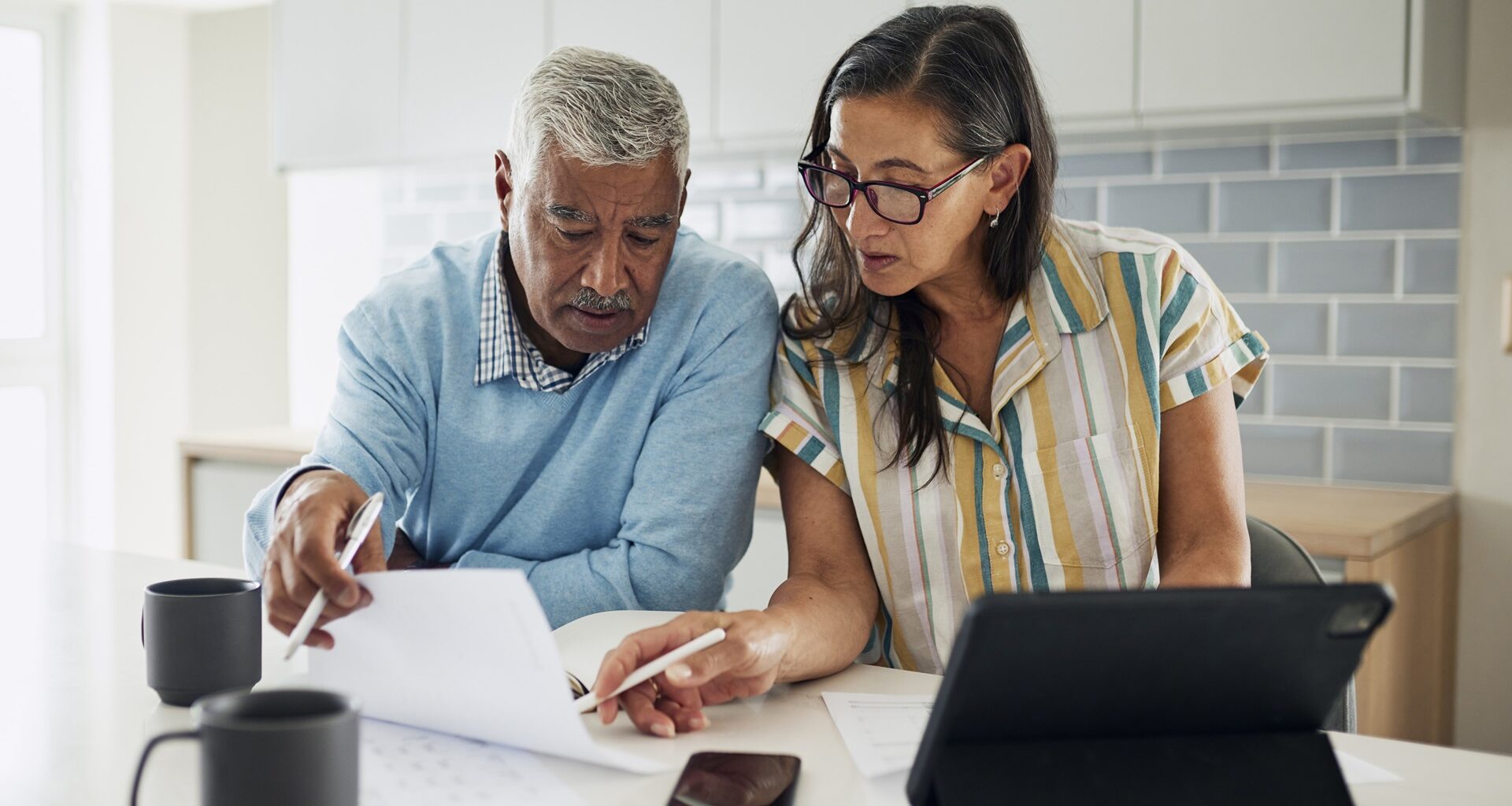 Shot of a senior couple standing in their kitchen going over finances on paper and on a tablet
