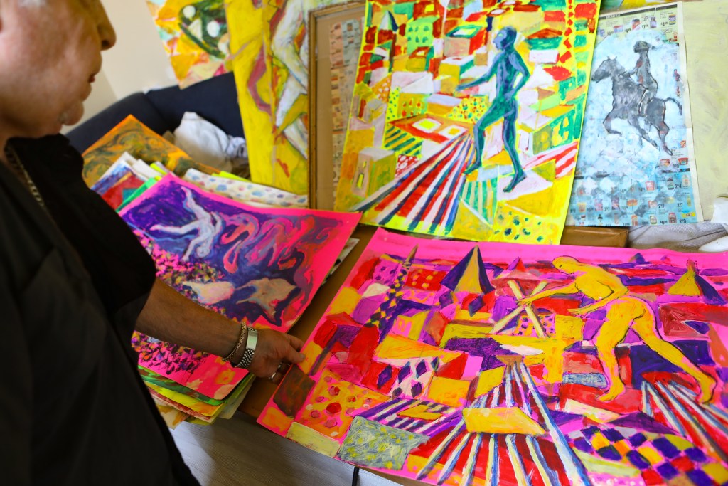 Artist Peter Passuntino is in the foreground wearing a black button down shirt and silver watch. His hand holds a pink painting with a figure in a theatrical city landscape.