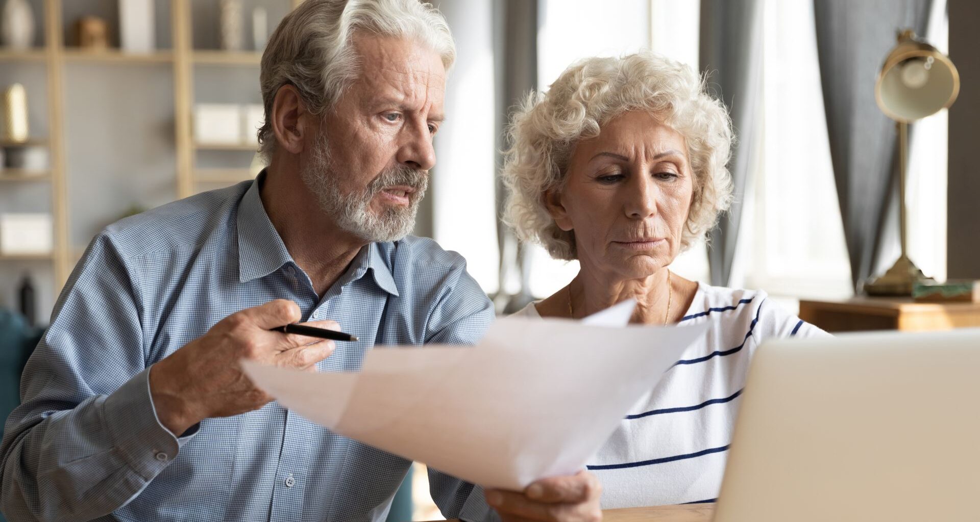 Stressed elderly married couple sitting at table at home manage budget paying using on-line banking access analyzing finance papers, having financial problems, high taxes expenses, unpaid debt concept.