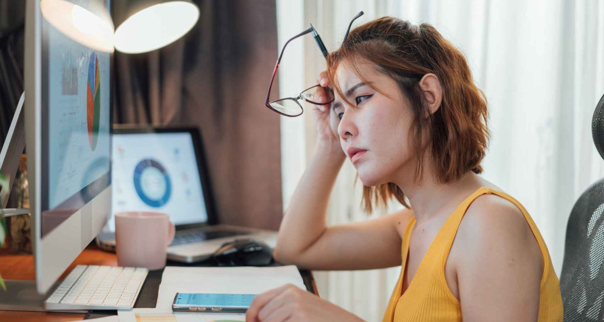 Stressed woman sitting at her desk, looking at a computer with financial charts on the screen.