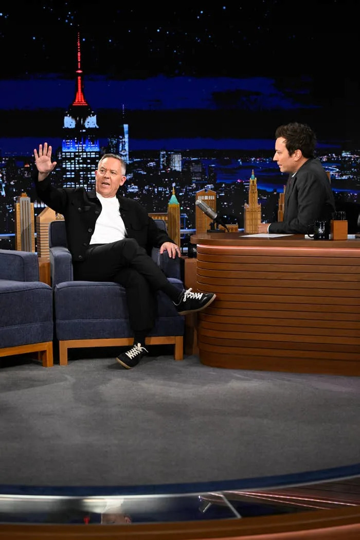 Greg Gutfeld on The Tonight Show waves while sitting on a sofa opposite the host at a desk with a cityscape backdrop