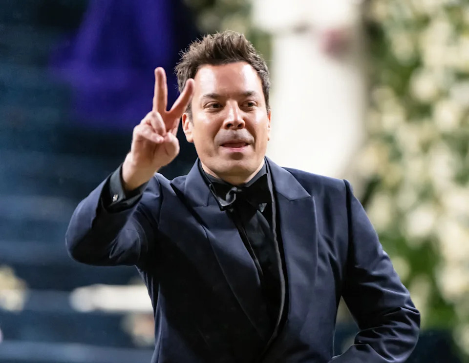 Jimmy Fallon in a formal suit flashes a peace sign at an event, with floral decorations in the background