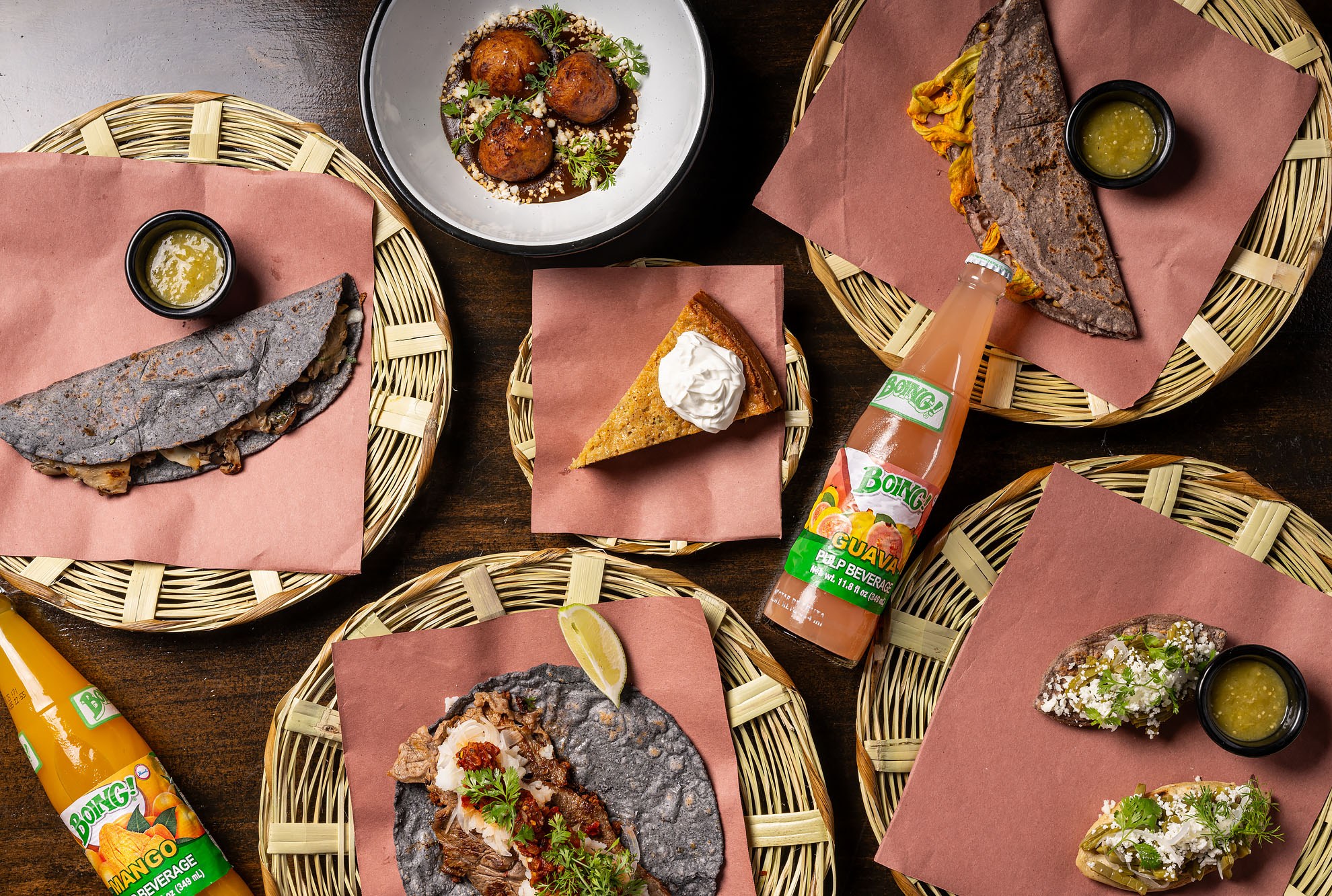 Overhead shot of a spread of dishes on a wood table with bottles of Boing at Komal Molino