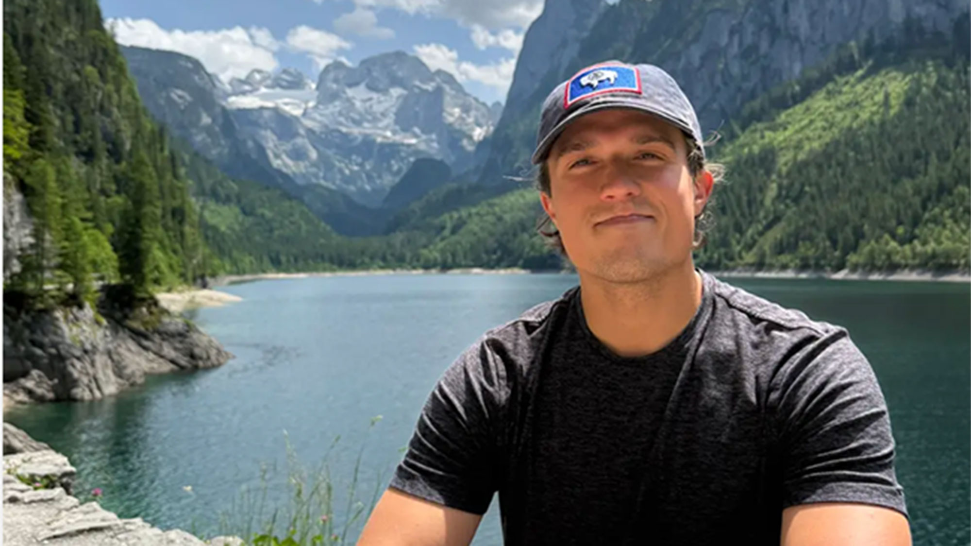 A handsome white man in his 20s, baseball cap and t shirt smiles for the camera in front of an alpine lake with mountains all around and blue sky with clouds in the background.