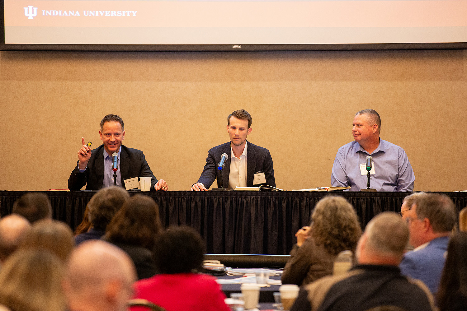 Three men - one in a dark suit, one in a dark sports coat, one in a blue shirt - talk during a panel discussion