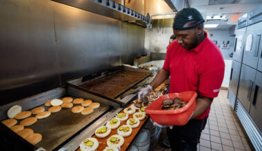 A cook in a red shirt and cap prepares burgers in a commercial kitchen, placing cooked patties on buns lined up on a counter.