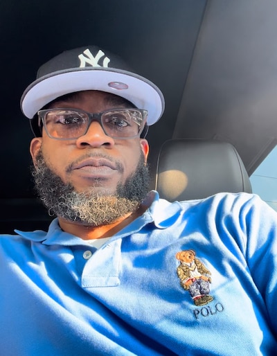 A photograph of a Black man wearing a blue dress shirt and a baseball hat poses for a selfie in a car.