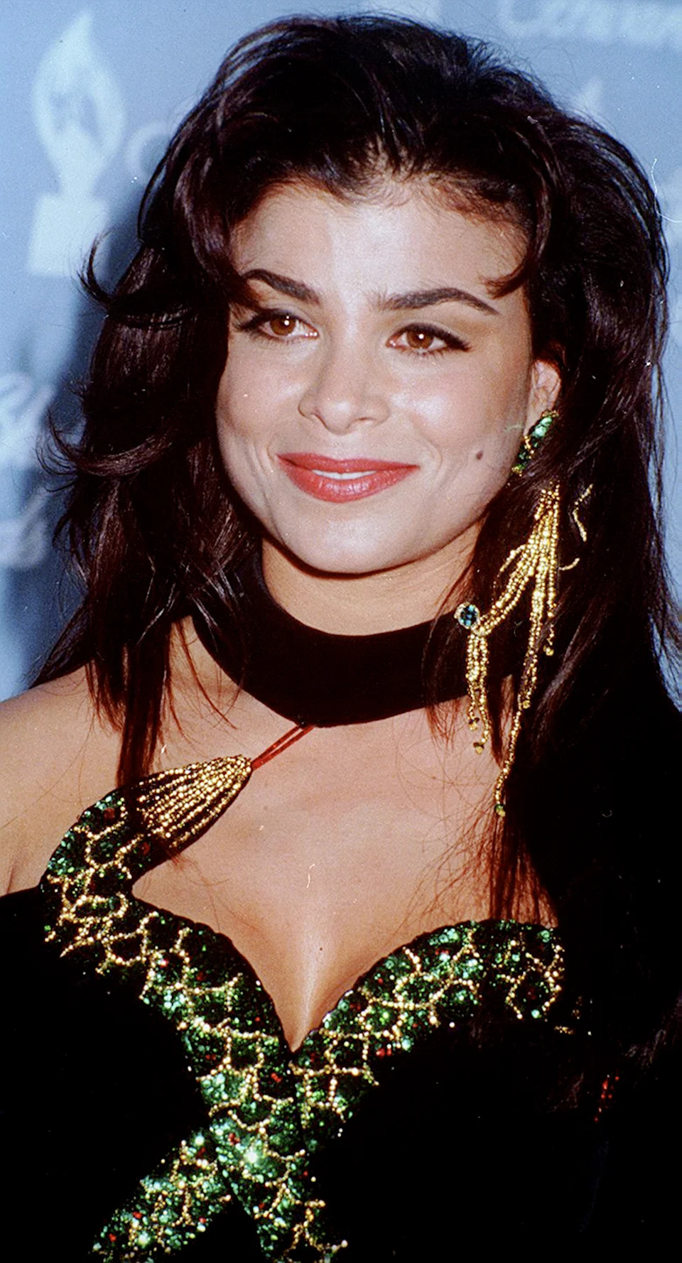 Person on red carpet in glamorous dress with gold and green sequin detailing, wearing statement earrings, smiling at an event