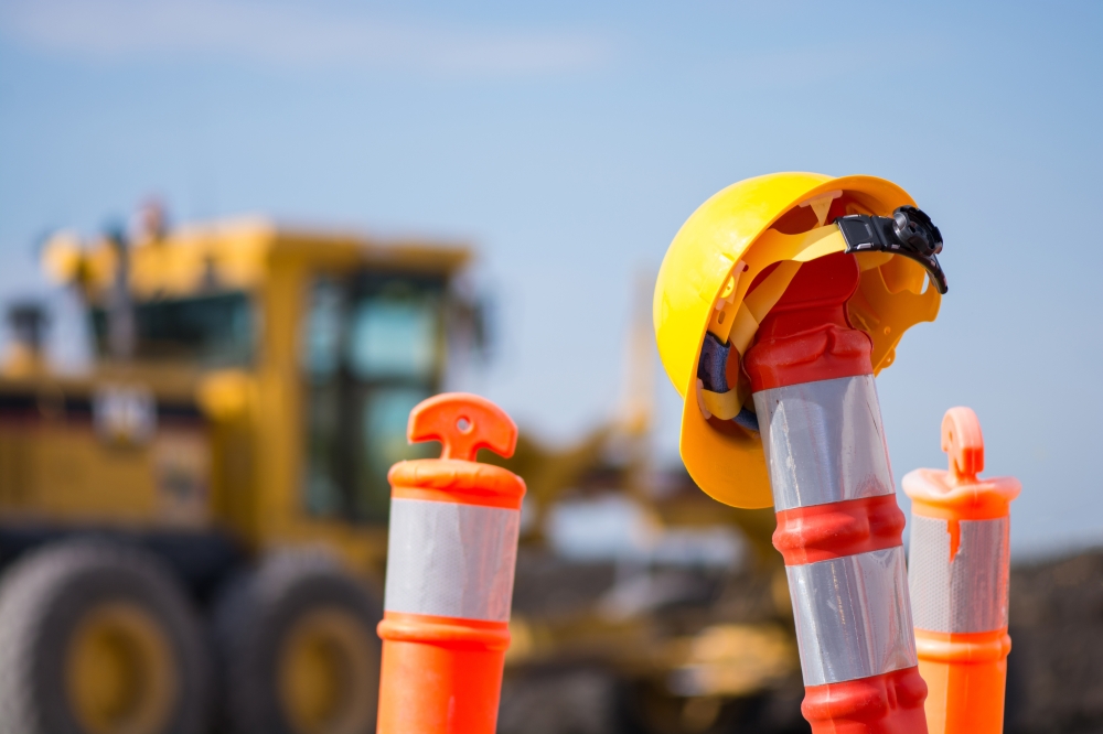 Thin road work cones with hard hat on top and construction machinery in background