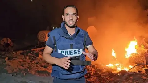 Al Jazeera Anas al-Sharif is wearing a blue flak jacket with 'PRESS' written across the front, and he's standing in front of burning debris.