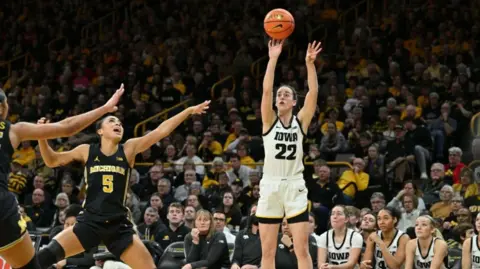 Reuters WNBA basketball player Caitlin Clark shoots a shot in a packed stadium, while other players try to block her shot. 