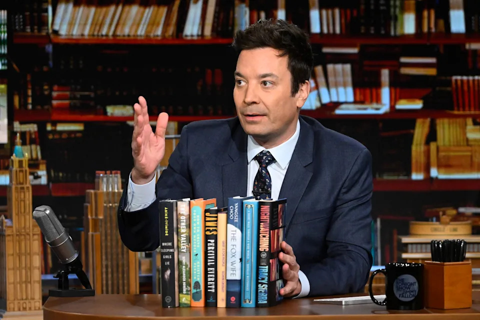 Jimmy Fallon in a suit seated behind a talk show desk discussing a selection of books, gesturing with one hand