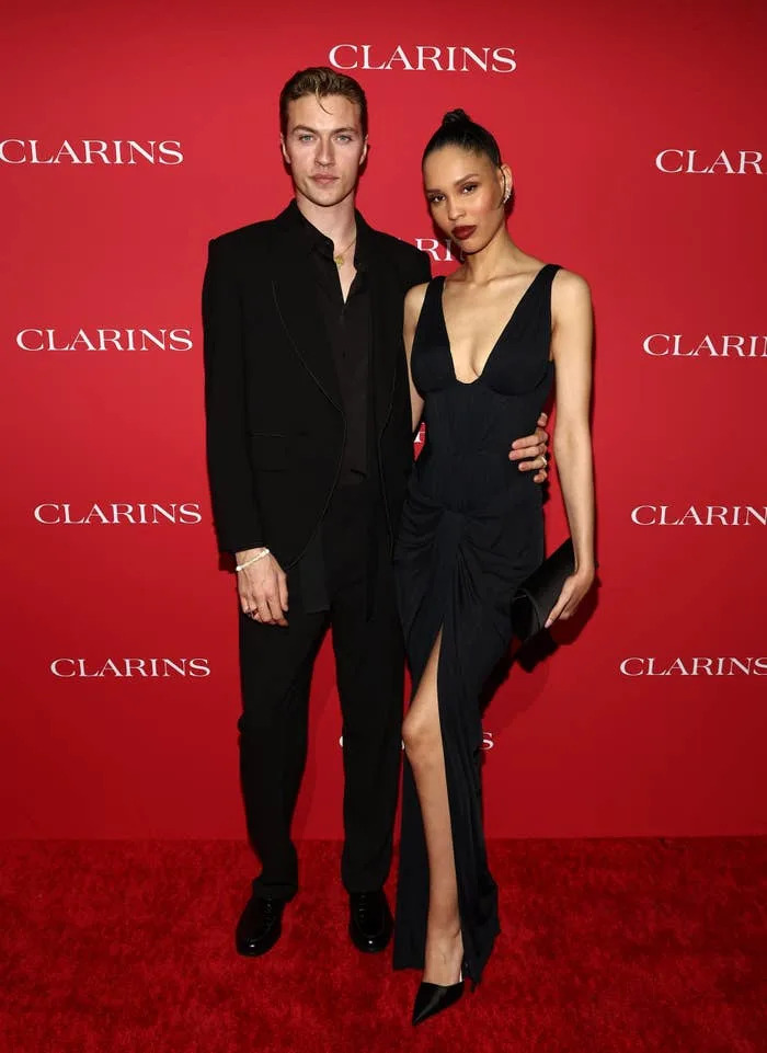 Lucky Blue Smith and Nara Smith posing on a red carpet. One is wearing a dark suit, the other a sleeveless gown with a slit. Red backdrop with "Clarins" logos