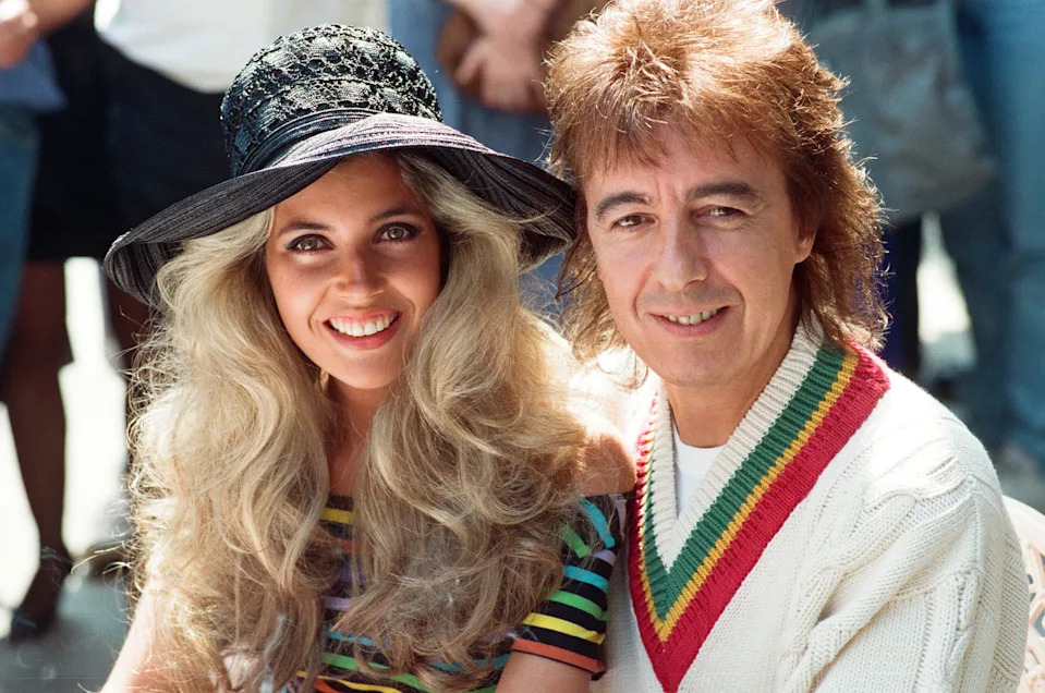 Two people smiling, woman in a striped top and large hat, man in a sweater with a colorful collar. 
