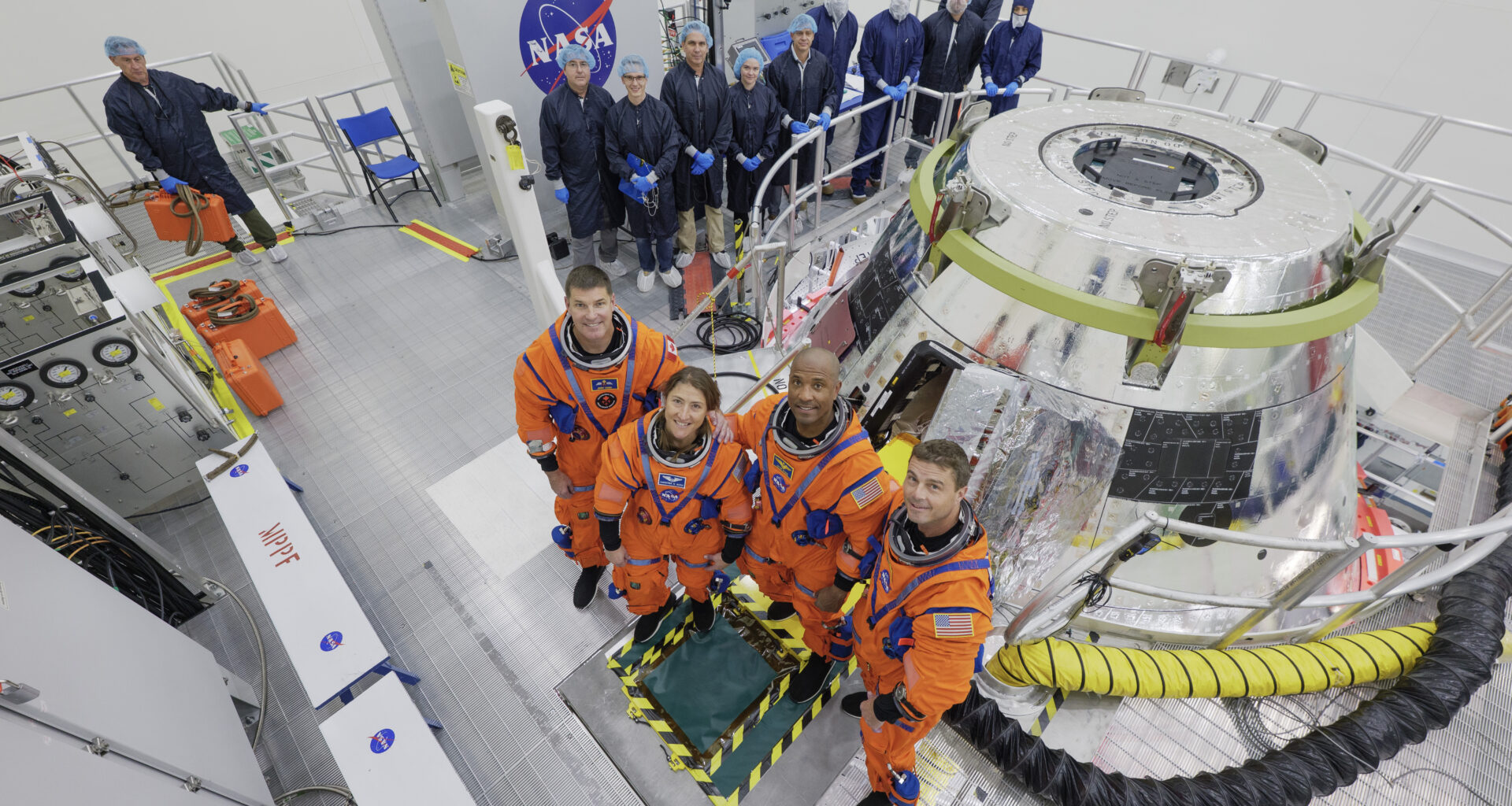 The Artemis II crew stands in front of the Orion spacecraft. They are wearing orange spacesuits without helmets. Orion is silver-colored and reflective. There is a round railing surrounding the spacecraft. There is also a group of people wearing blue jumpsuits, hairnets, and gloves standing behind Orion; they are members of the Artemis II closeout crew.