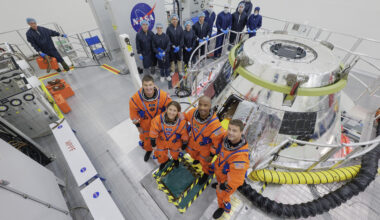 The Artemis II crew stands in front of the Orion spacecraft. They are wearing orange spacesuits without helmets. Orion is silver-colored and reflective. There is a round railing surrounding the spacecraft. There is also a group of people wearing blue jumpsuits, hairnets, and gloves standing behind Orion; they are members of the Artemis II closeout crew.