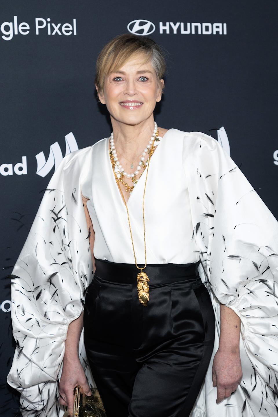 Person in elegant white blouse with voluminous sleeves and black pants, accessorized with layered necklaces, standing at a celebrity event