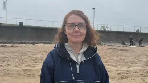 Norma Rae standing on the beach. She has shoulder length light brown hair, wearing glasses, a blue raincoat and grey half zip underneath.